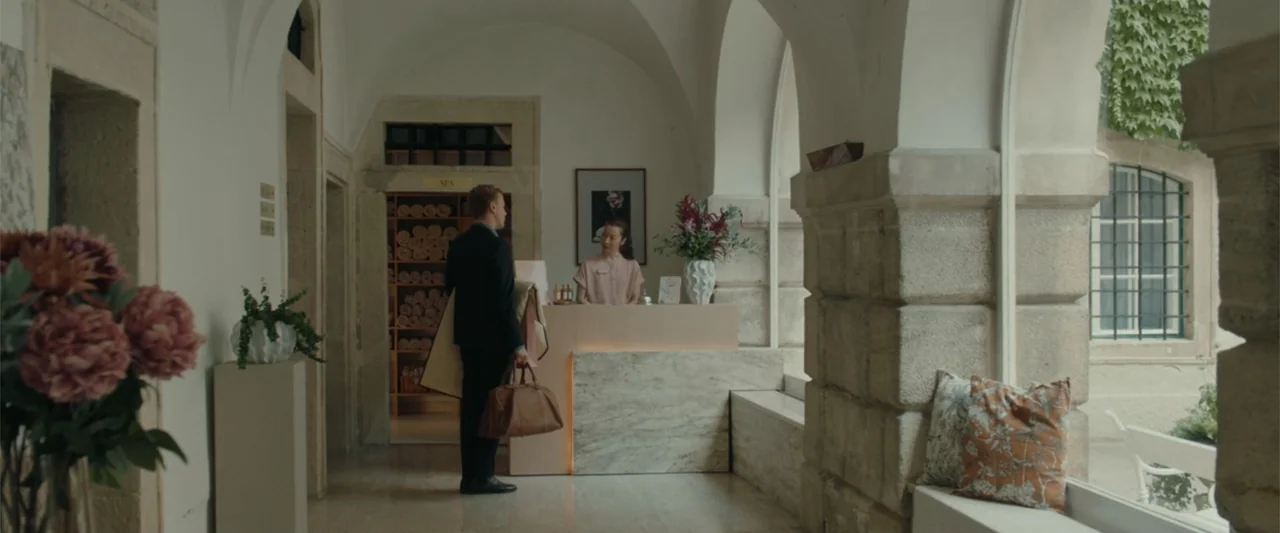 Peacock (2010) movie still: medium — A hotel reception area with stone arches and a marble-faced front desk. A man in a suit approaches the desk…