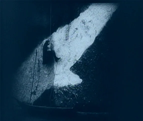 Destiny (1921) movie still: wide — A sheer dark rock formation dominates the frame against a black background. A lone figure rappels down the side…
