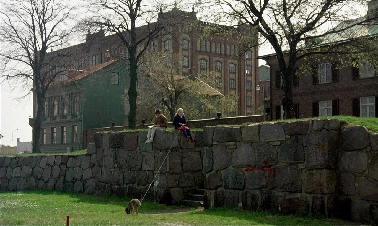 My Life As A Dog (1985) movie still: wide — A field borders a low, rough stone wall in front of old brick buildings. Two children sit on the wall…