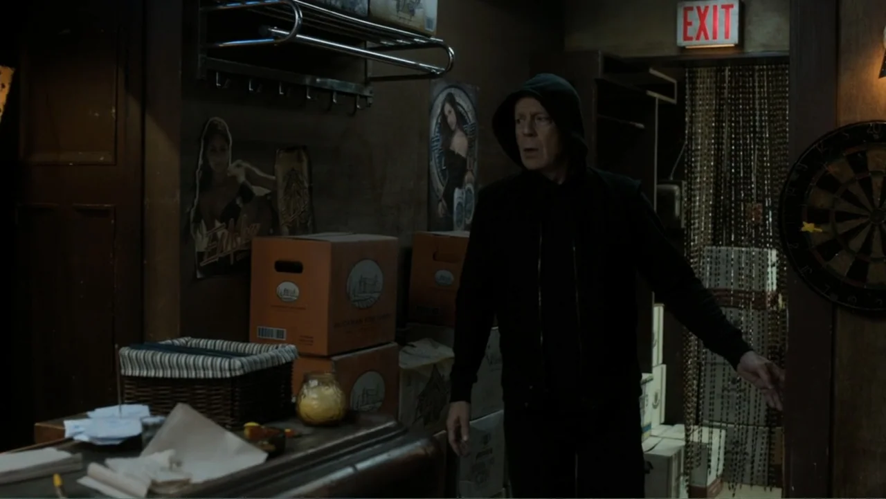Death Wish (2018) movie still: medium — A cluttered back room features stacked cardboard boxes and a dartboard on a wooden wall. An older man…