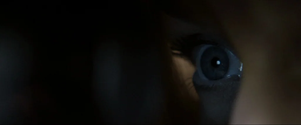 Clown (2014) movie still: closeup — A sliver of light reveals a single eye peeking through a dark, narrow crevice. The surrounding area is obscured…