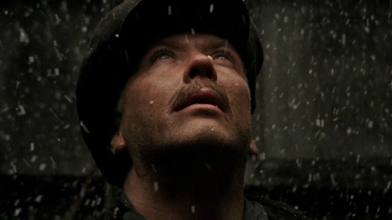 Everlasting Moments (2008) movie still: closeup — A man in a dark cap stands outdoors during a night rainstorm. He gazes directly upward while rain…