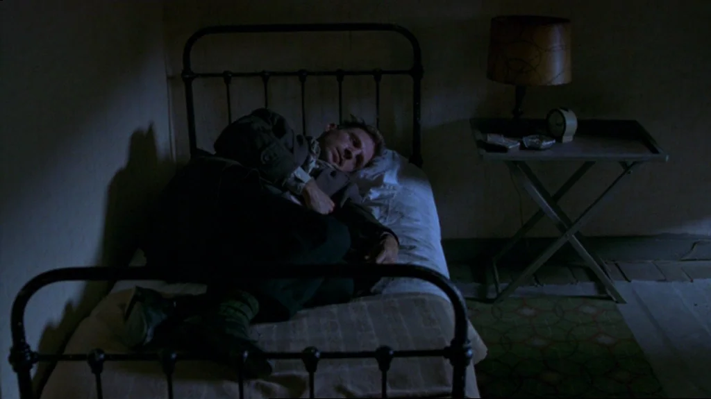 Spider (2002) movie still: medium — A dimly lit, sparse room contains a simple metal bed and a side table. A man rests on the bed while fully…