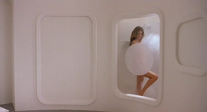 CQ (2001) movie still: medium — A minimalist white room features rounded recessed wall compartments typical of space-age design. A woman stands in…