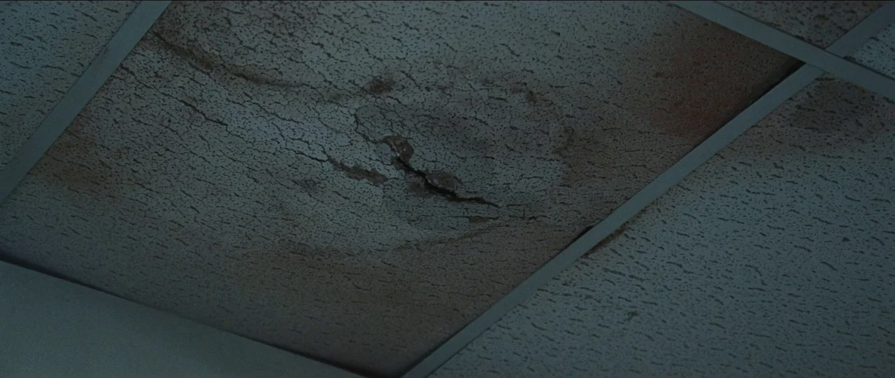 Bad Education (2019) movie still: closeup — A drop ceiling features a square tile with a dark water-stained crack. The porous material shows signs…