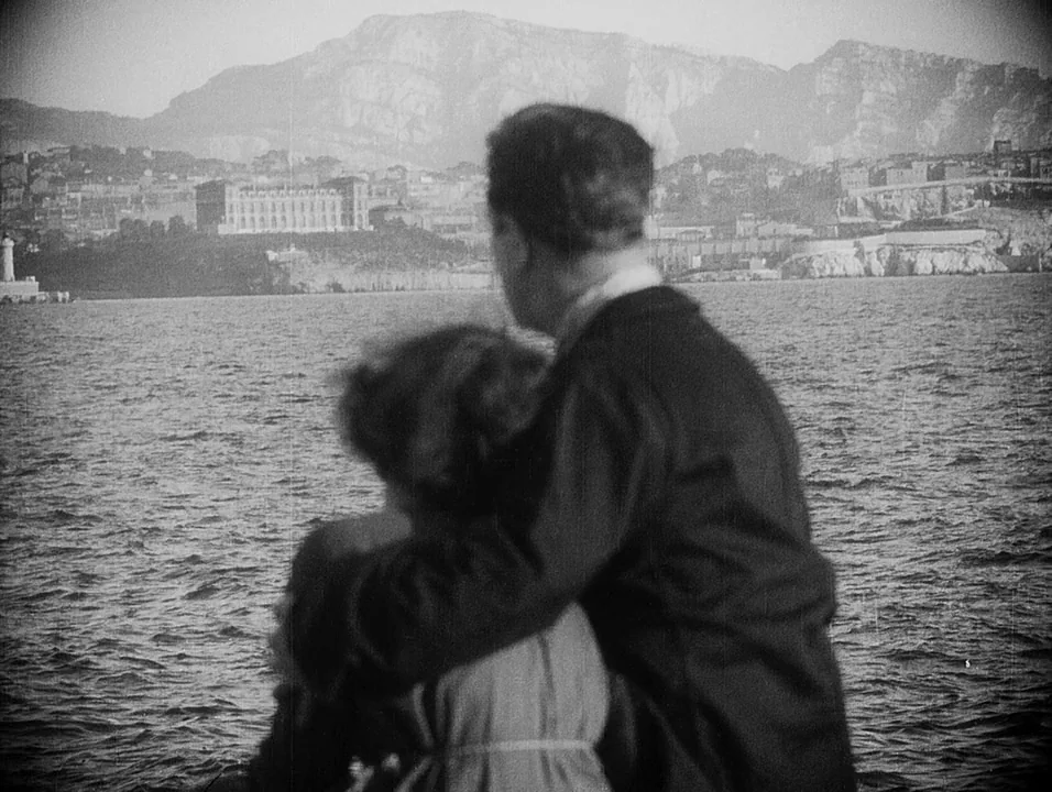 Coeur Fidele (1923) movie still: medium — A man and woman viewed from behind on a boat facing a distant rocky coastline. Large mountains rise in the…