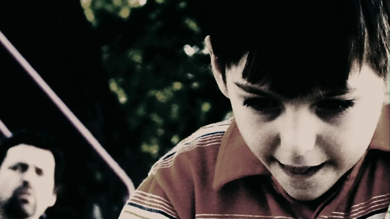 Saw III (2006) movie still: closeup — A close-up shot of a young boy against an outdoor playground structure. A blurred man stands in the background…