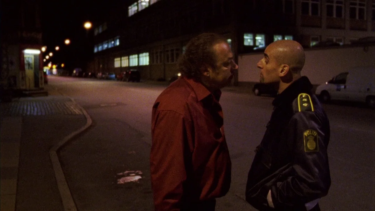 Pusher III (2004) movie still: medium — An empty urban road at night features a modern building with lit windows. A middle-aged man in a red shirt…