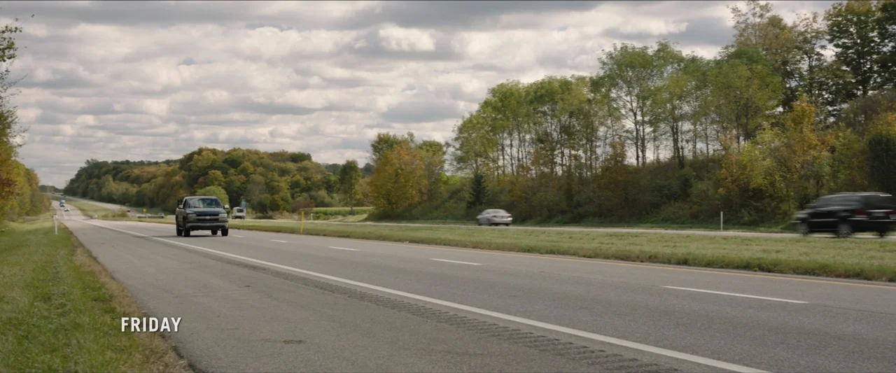 The Marksman (2021) movie still: wide — A multi-lane asphalt highway stretches through a rural landscape with dense trees. Several passenger…