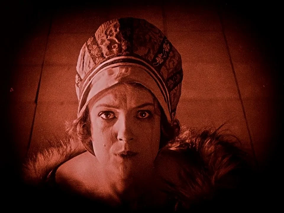 Häxan (1922) movie still: closeup — A close-up shot of a woman in an ornate cap against a dark background. She looks directly toward the camera with…