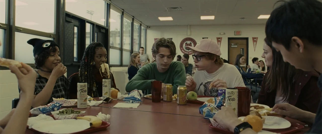 Chemical Hearts (2020) movie still: medium — A high school cafeteria features rows of red tables and large windows. Several teenagers sit together…