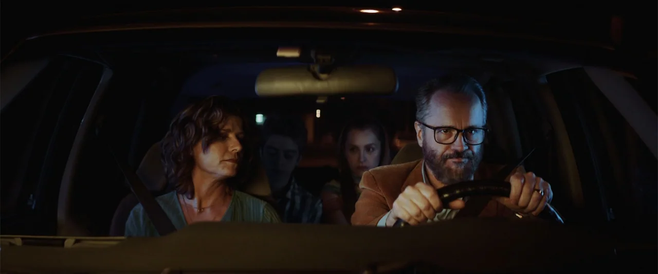 The Boy Who Killed My Parents (2021) movie still: medium — Inside a moving vehicle at night, a family of four sits in the seats. The driver keeps…