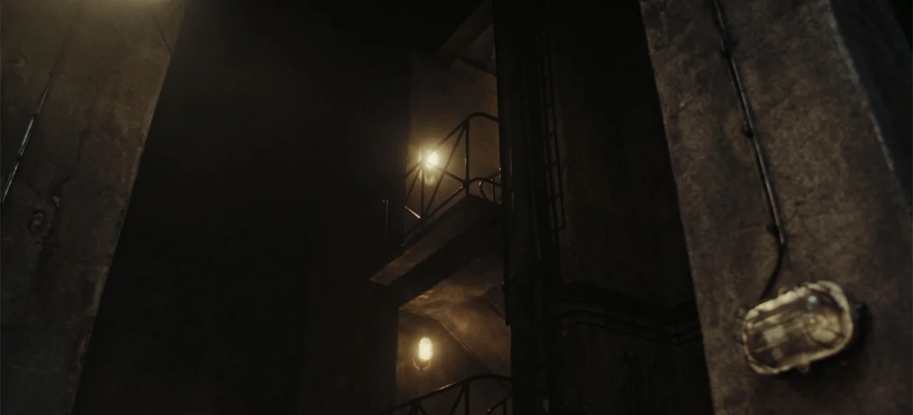 The Tank (2025) movie still: establishing — A dark concrete industrial stairwell features metal railings and dim wall-mounted practical lights. The…