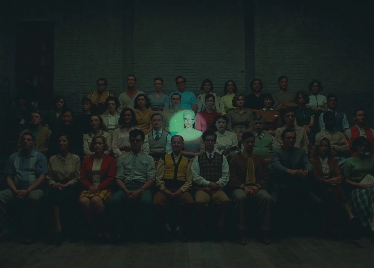 Asteroid City (2023) movie still: wide — A large crowd of people sits in rows inside a dark, industrial brick building. One woman in the center is…