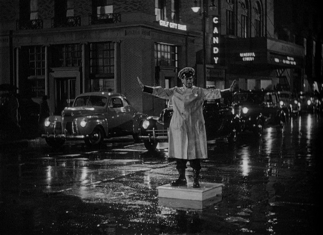 Dead Reckoning (1946) movie still: fullbody — A wet paved street at night with parked vintage cars and city building facades. A traffic officer…
