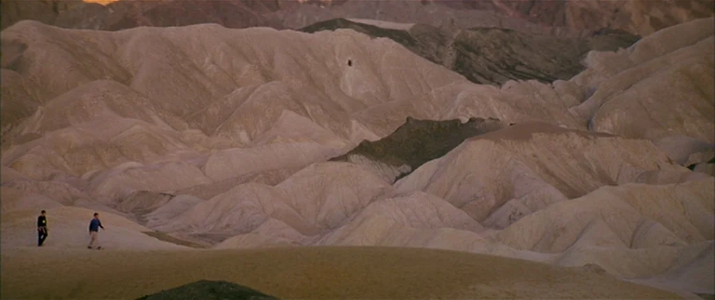 Gerry (2002) movie still: wide — Extensive rolling hills of beige, eroded dirt and rock form a vast desert landscape. Two small figures stand on a…