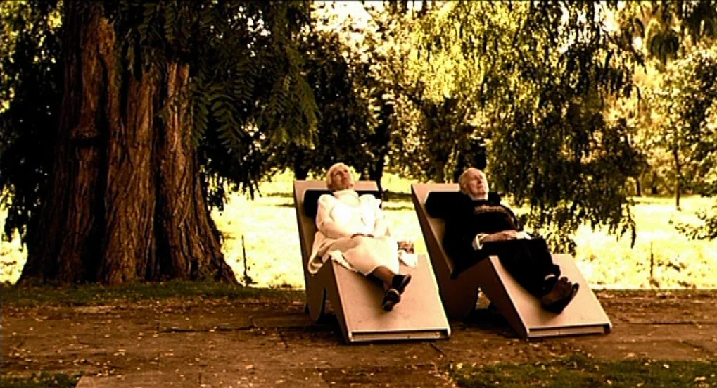 Jeux d’Enfants (2003) movie still: fullbody — An elderly couple reclines in minimalist lounge chairs set on a paved park area. A large tree stands…