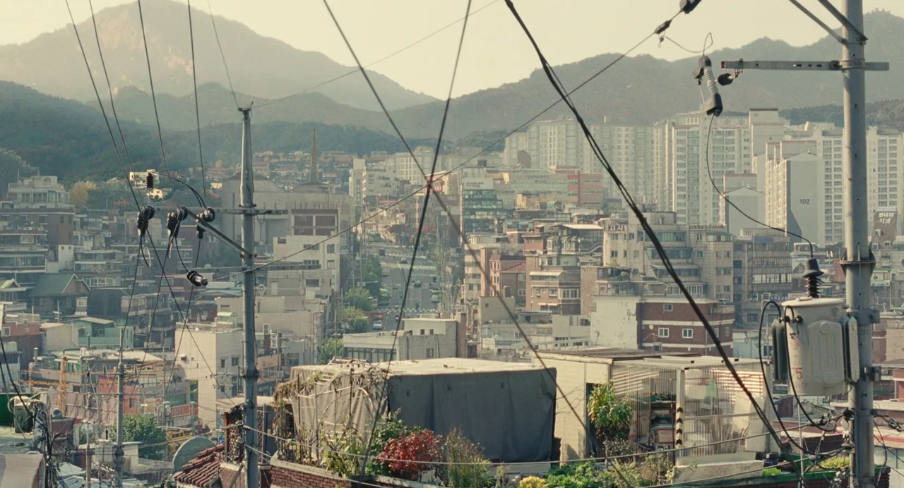 Past Lives (2023) movie still: establishing — A dense urban neighborhood sits beneath rolling mountains during a bright clear day. Tangled power…