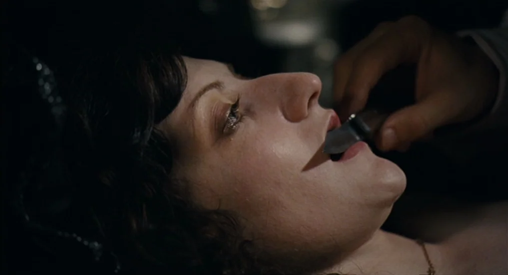 House of Tolerance (2011) movie still: closeup — A dark, dimly lit room where a woman lies down. A man's hand holds the tip of a small knife against…