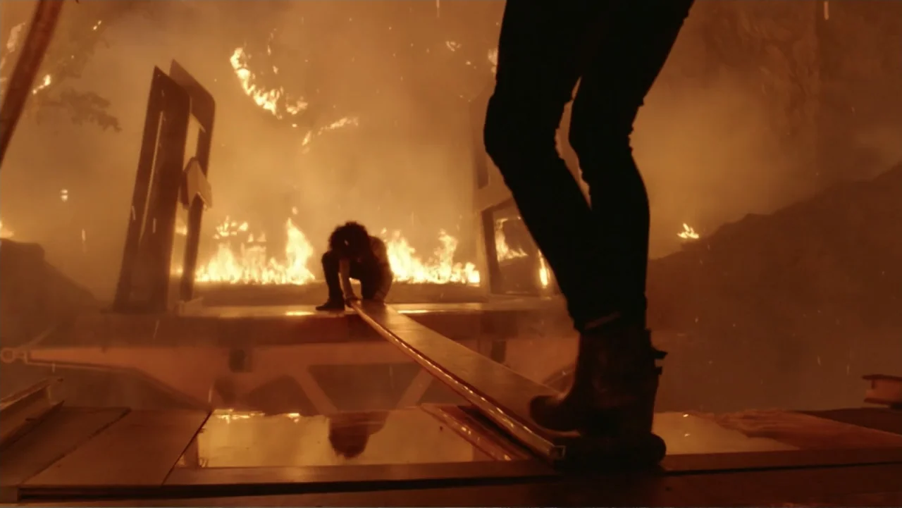 Skyscraper (2018) movie still: medium — A person walks across a narrow metal beam at a burning site surrounded by debris. A second person crouches…