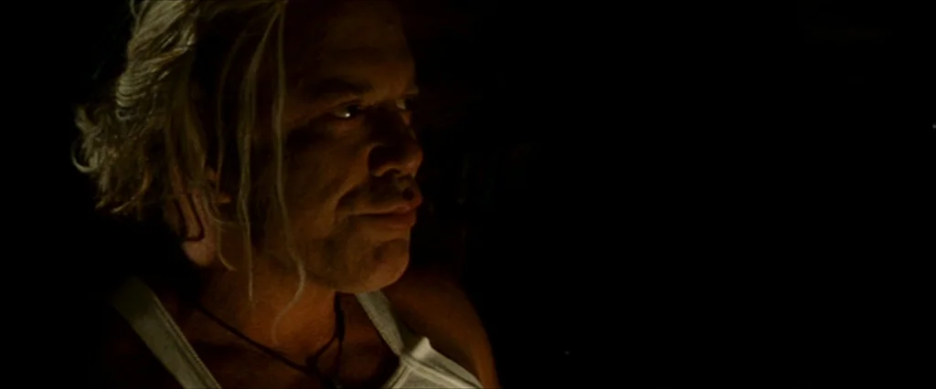 The Wrestler (2008) movie still: closeup — A dark, underexposed interior space surrounds a man with shoulder-length hair. The subject looks…
