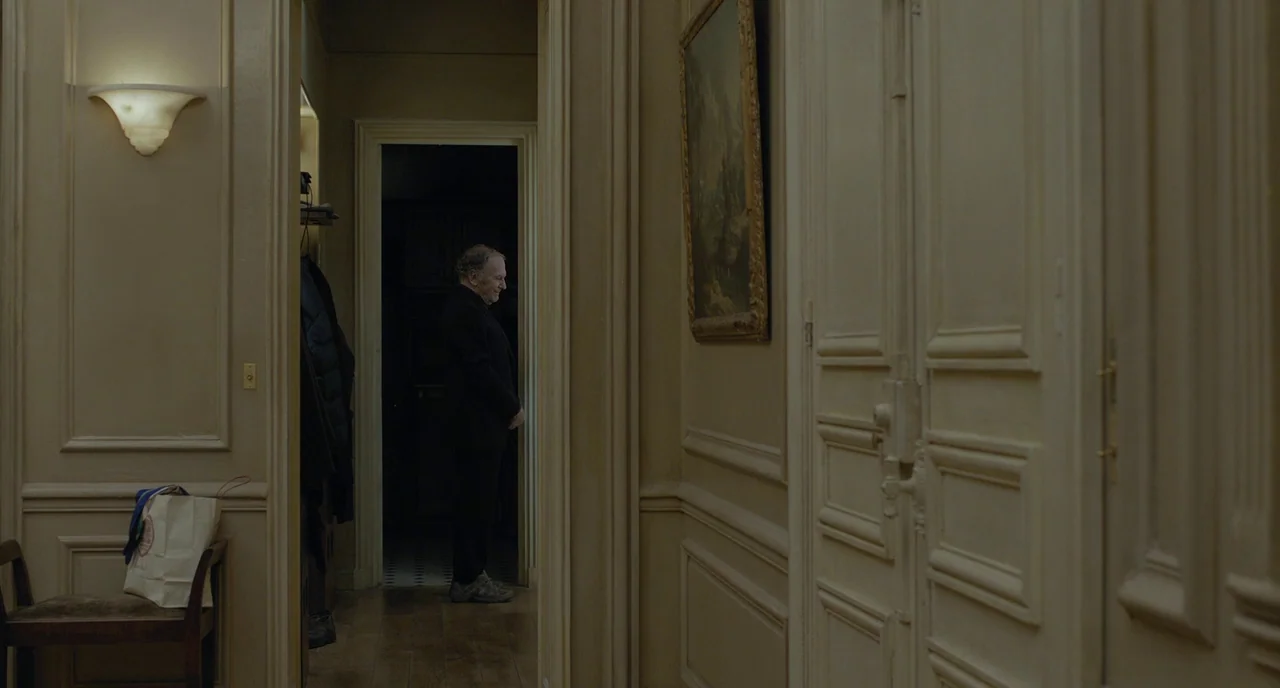 Amour (2012) movie still: wide — A classic residential corridor features paneled walls painted in shades of beige and cream. A man wearing a black…