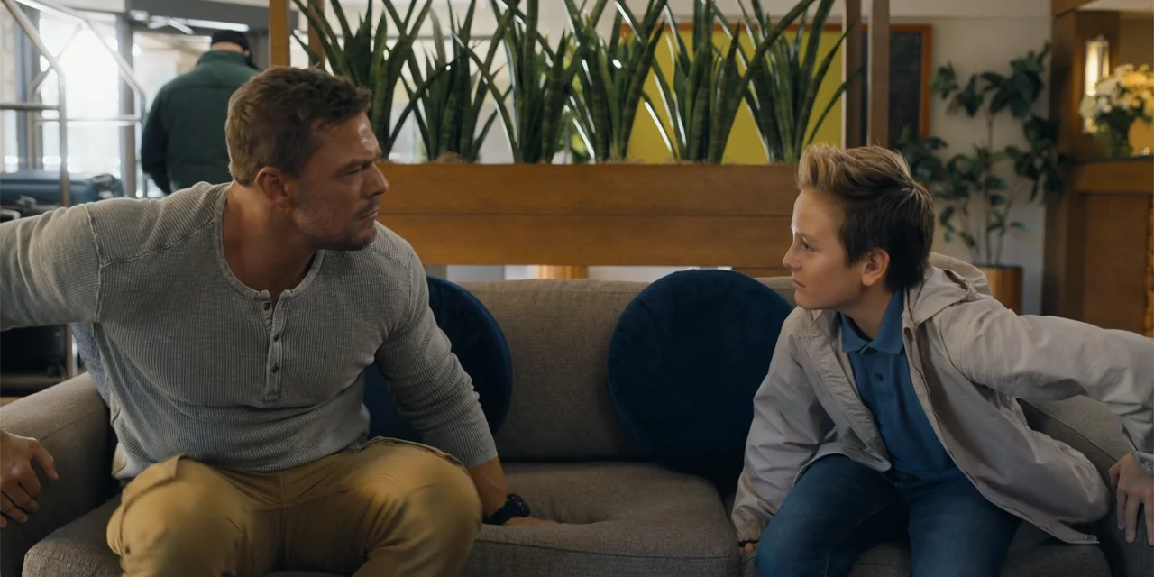 Playdate (2025) movie still: medium — A hotel lobby features a large grey sofa and tall indoor plants behind it. A man and a teenage boy are seated…