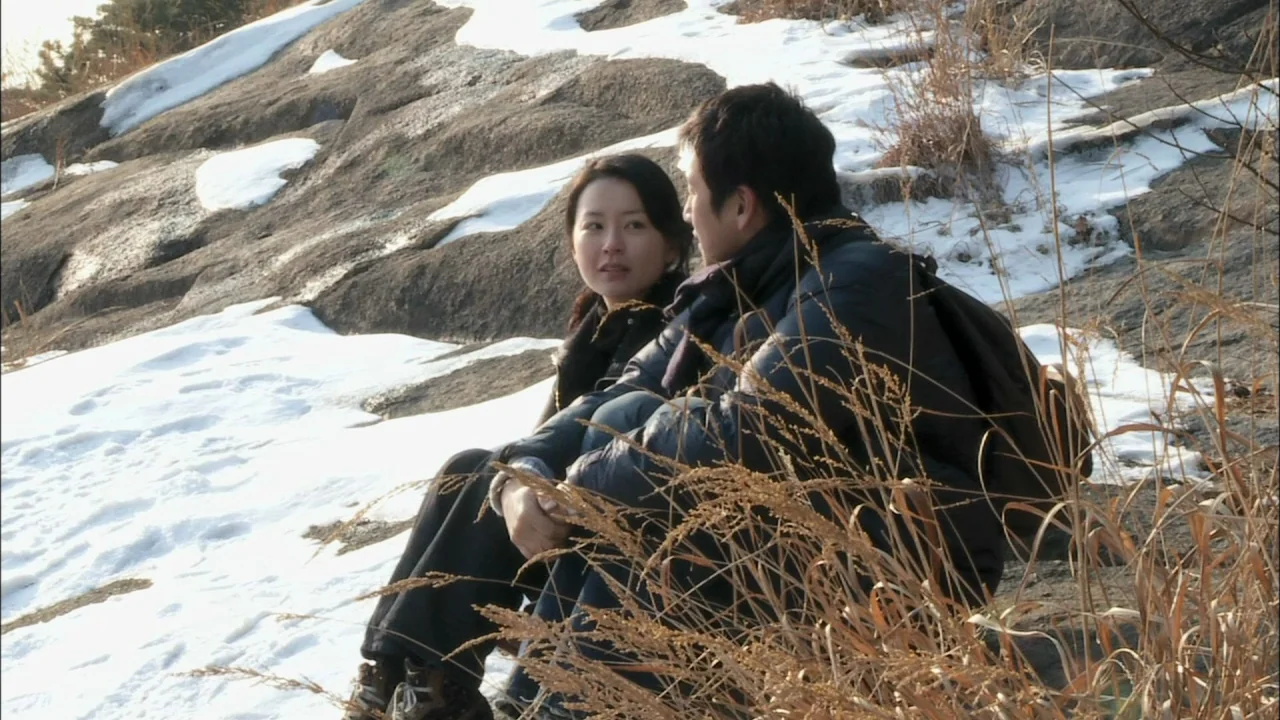 Oki’s Movie (2010) movie still: medium — A rocky incline features patches of melting snow and dried, tall golden grass. A man and woman sit closely…