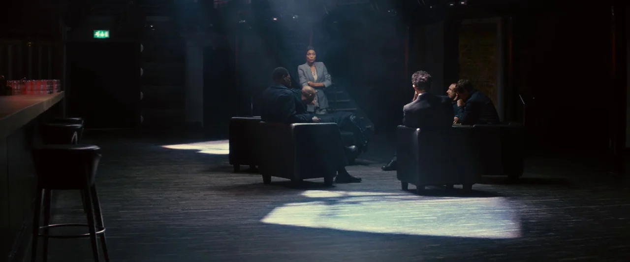 Trance (2013) movie still: wide — A dark, expansive room contains a group of people clustered in armchairs and standing. Patches of intense light…