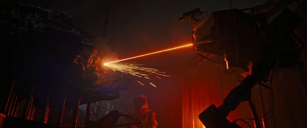 Alien: Romulus (2024) movie still: medium — A dark laboratory space features a mechanical arm projecting a laser onto a rock. A hooded person stands…
