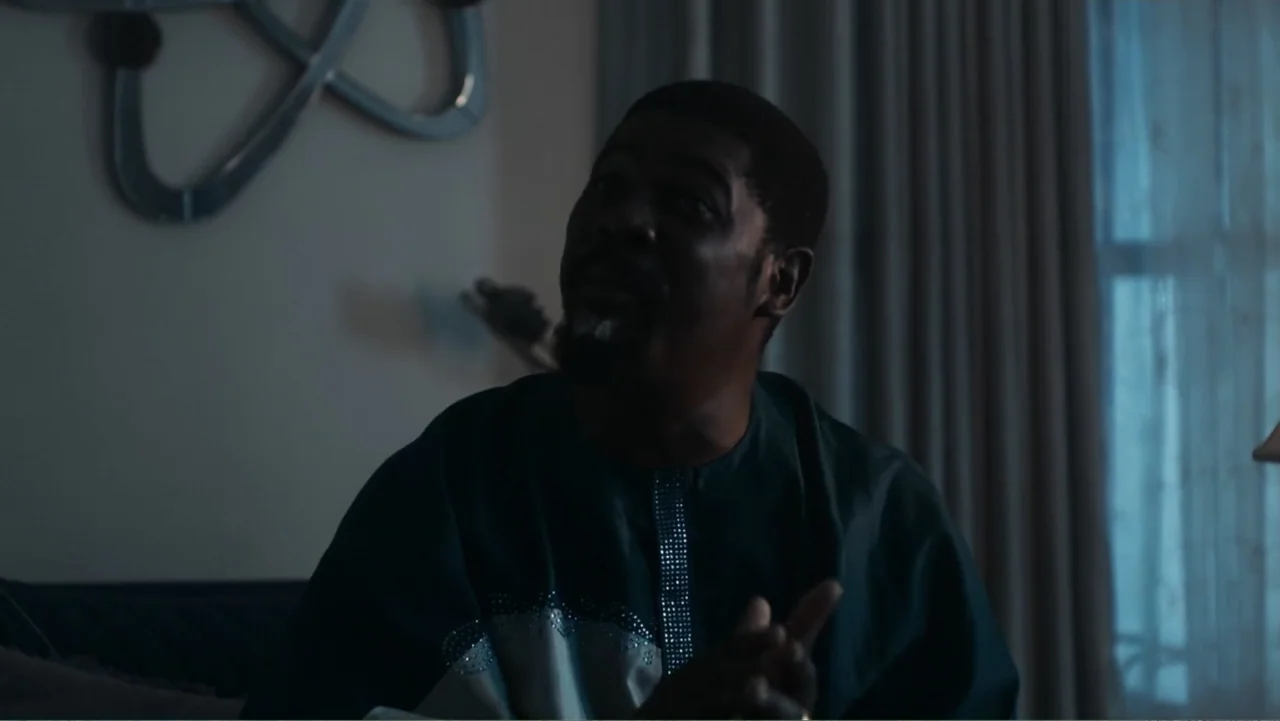 Gangs of Lagos (2023) movie still: medium — A dimly lit room with draped curtains visible in the background. A middle-aged man sits in a dark tunic…