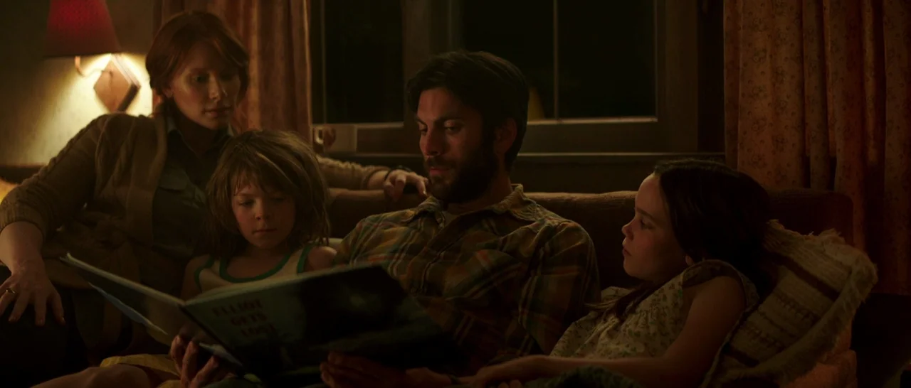 Pete’s Dragon (2016) movie still: medium — A dimly lit room with a couch against a dark window at night. A family of four sits together while the…