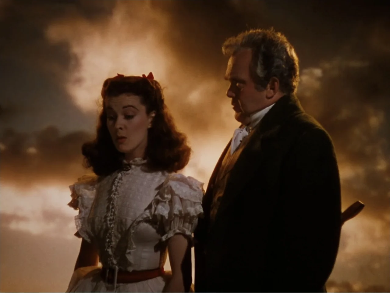 Gone with the Wind (1939) movie still: medium — Two people stand in an open outdoor setting against a vibrant, cloud-filled sunset sky. A young…