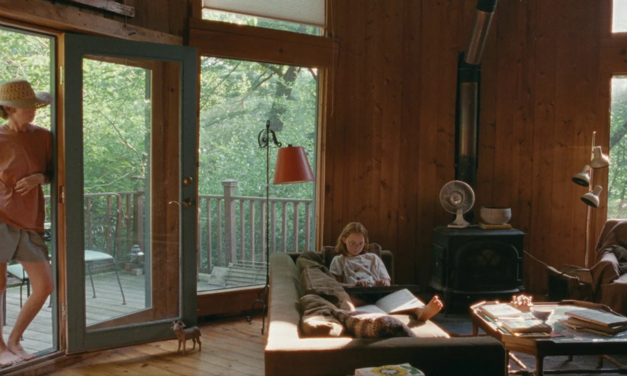 Janet Planet (2023) movie still: wide — A rustic cabin room has wood-paneled walls and large windows overlooking a green forest. One person stands…