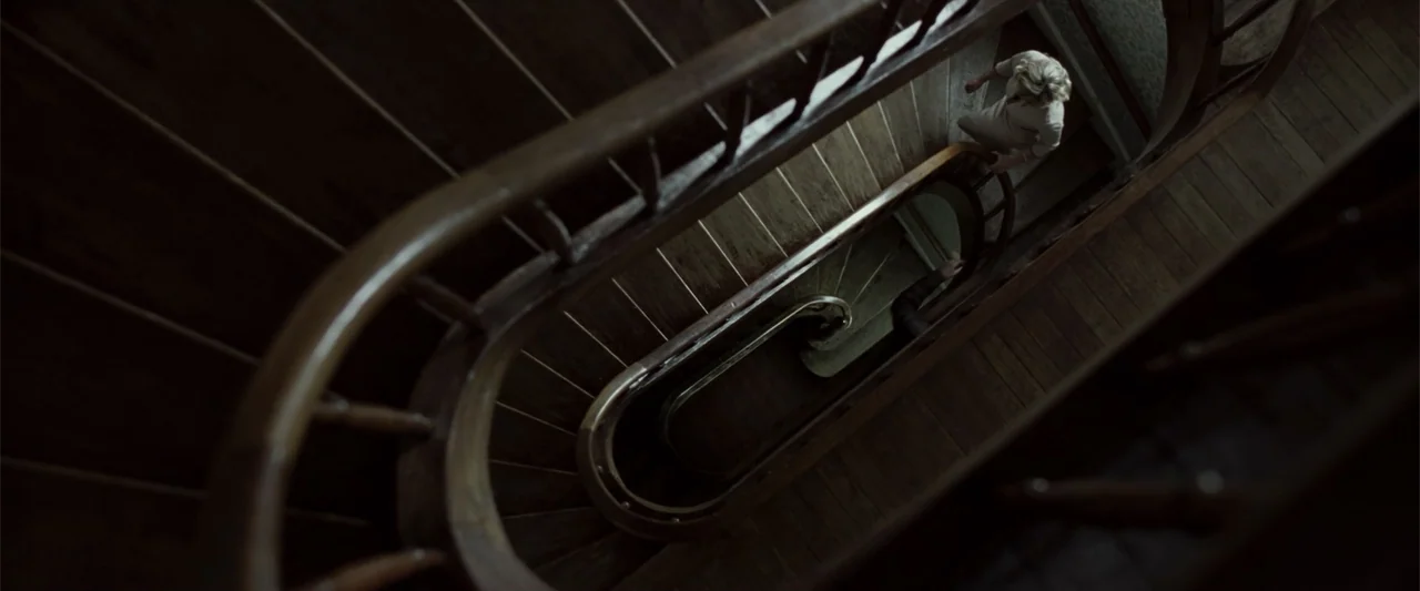 Anna (2013) movie still: wide — A wooden spiral staircase descends into deep shadow with a woman standing above. She looks down toward an obscured…