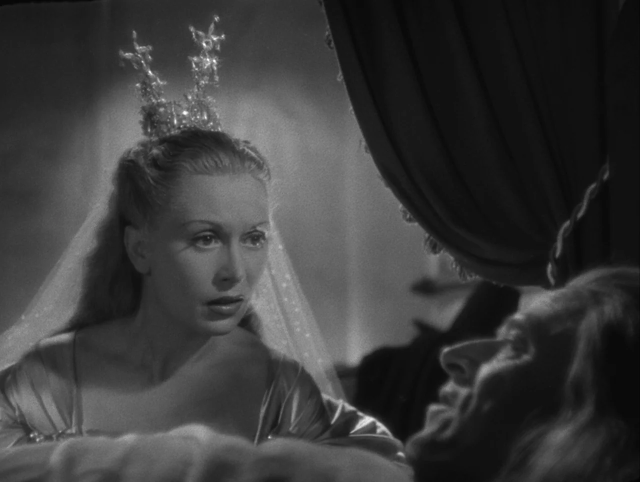 La Belle et la Bête (1946) movie still: medium — A dimly lit room features a woman in a crystalline crown facing a man. The man reclines on a bed…