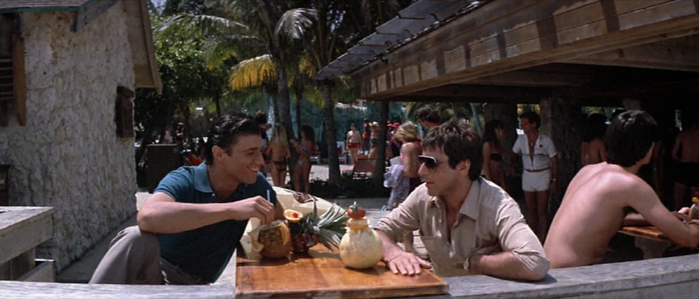 Scarface (1983) movie still: medium — An outdoor bar area features a wooden counter sheltered by a large roof structure. Two men sit at the counter…