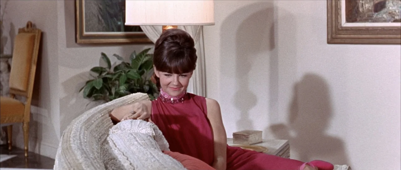 Clambake (1967) movie still: medium — A 1960s living room features a textured off-white sofa and a nearby floor lamp. A woman in a magenta dress…