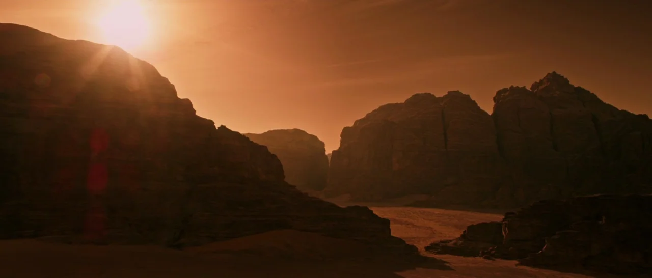 The Martian (2015) movie still: establishing — A vast rocky desert canyon spans the frame under a bright orange sun. Sand dunes sit between the…
