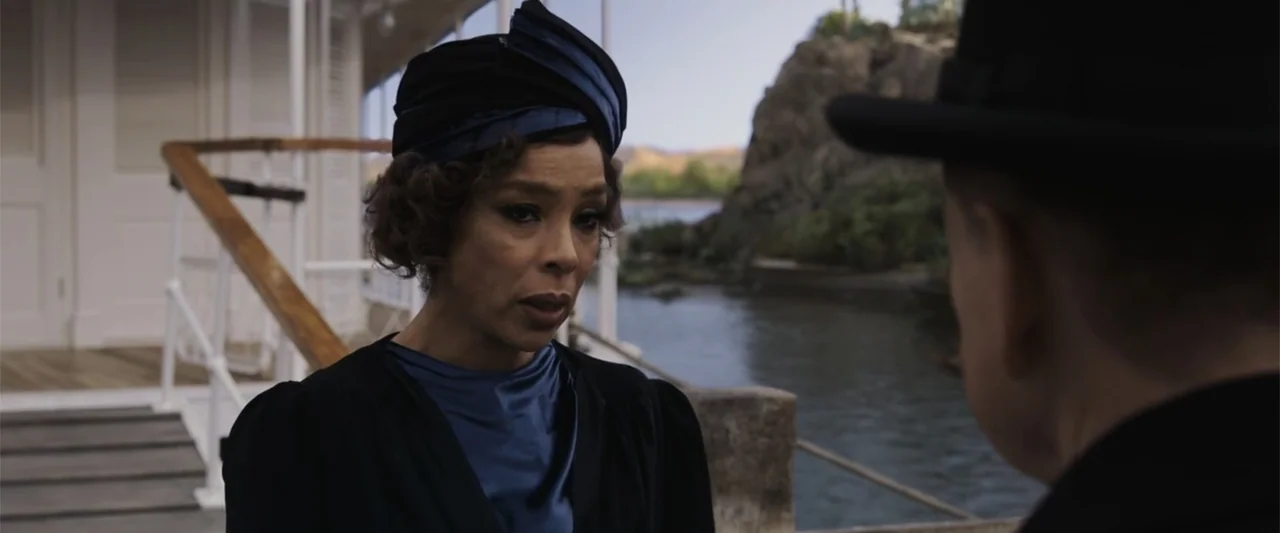 Death on the Nile (2022) movie still: medium — A woman stands on a wooden dock near a rocky shoreline during the day. She faces a man whose back is…