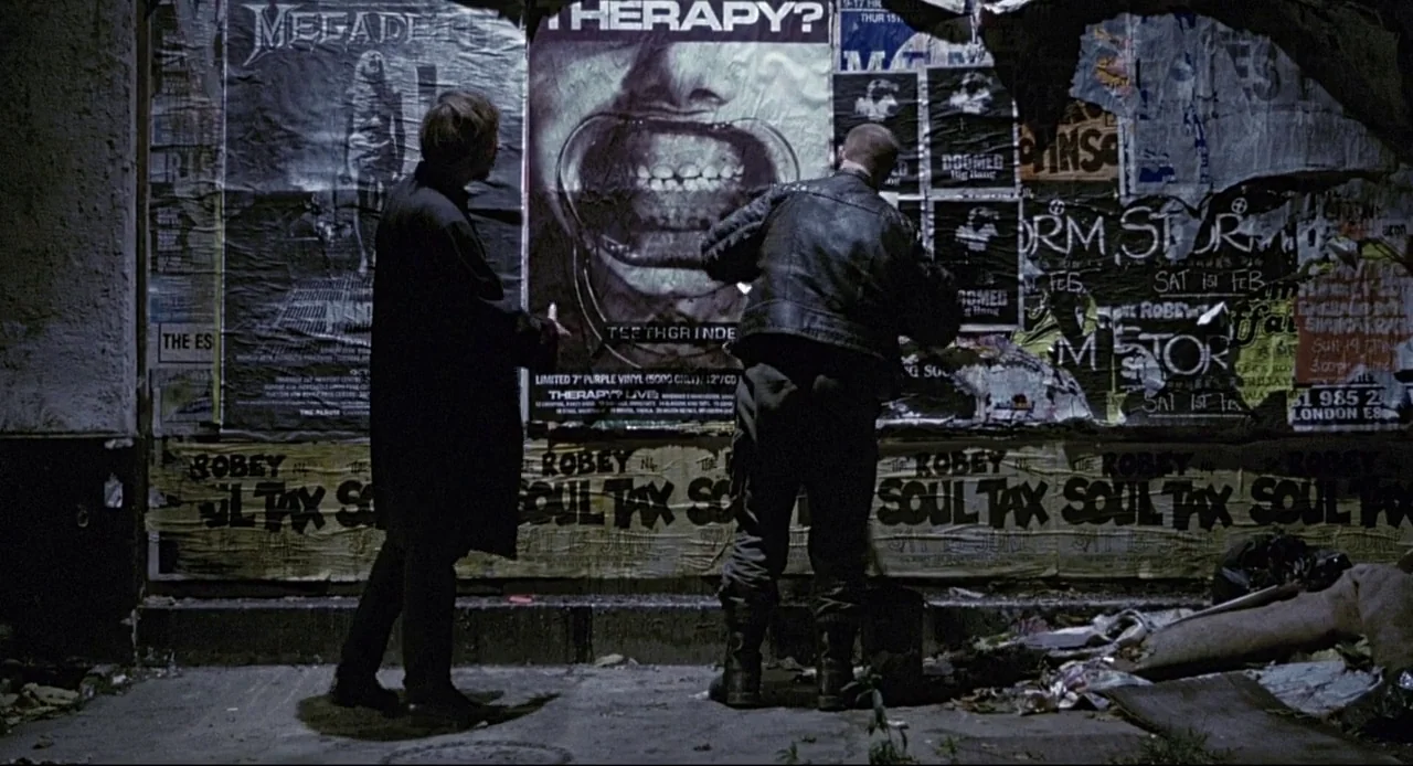 Naked (1993) movie still: fullbody — A dark city wall covered in overlapping concert posters and flyers from the 1990s. Two men stand in the dim…