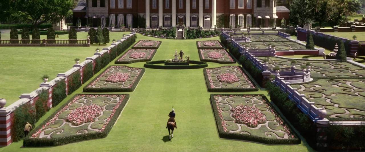 The Great Gatsby (2013) movie still: wide — A expansive manicured lawn features geometric floral beds and a distant brick mansion. A lone rider on a…