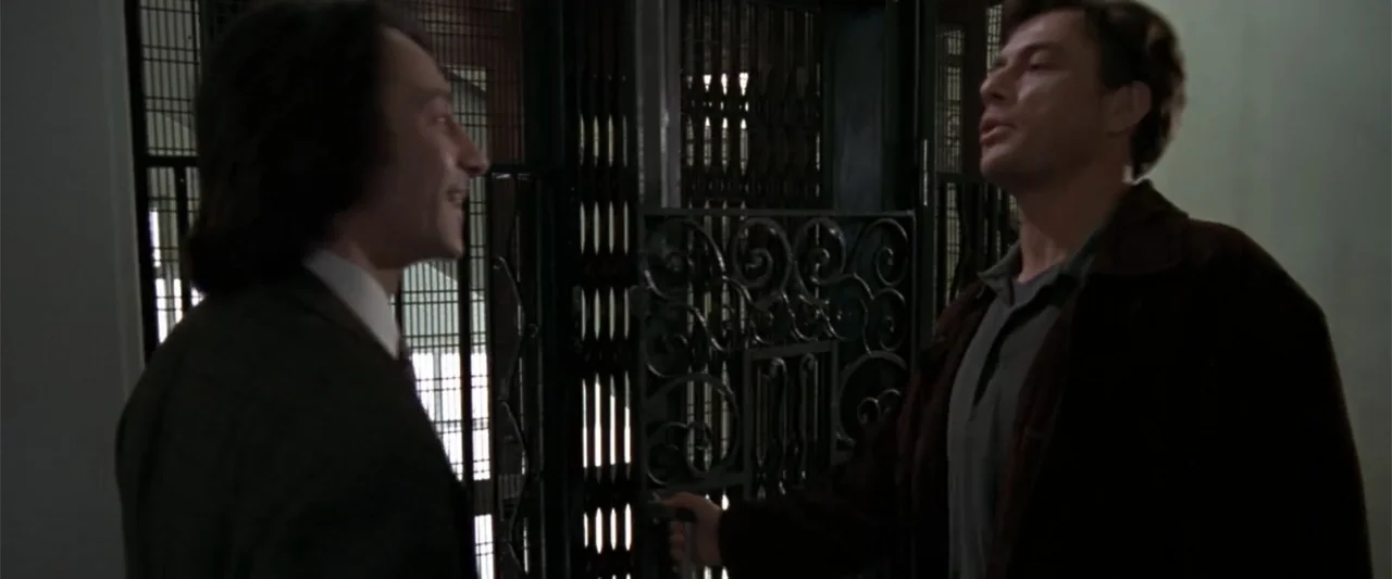 Maximum Risk (1996) movie still: medium — An elevator lobby with a dark ornamental metal gate and plain walls. Two men stand in profile to one…