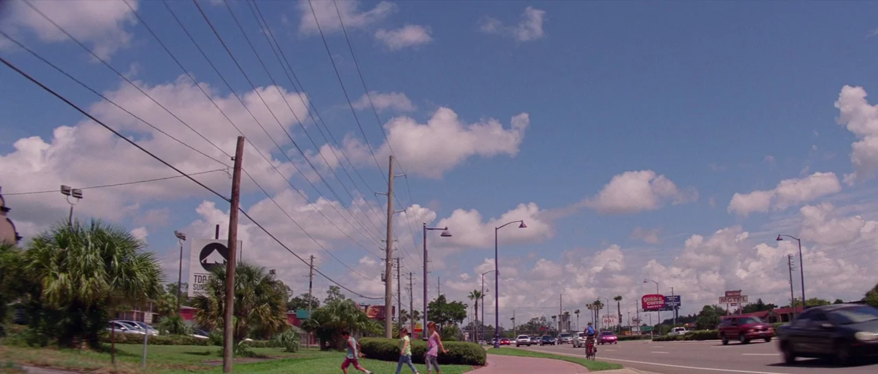 The Florida Project (2017) movie still: wide — A suburban roadside with power lines, palm trees, and various commercial signage overhead. Three…