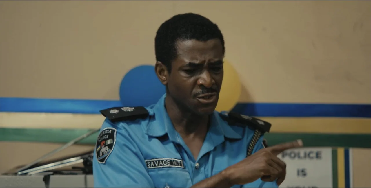 Project Assault (2025) movie still: medium — An interior police station office features a tan wall with blue and yellow decorative bands. A…