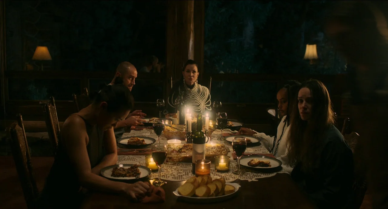 Trim Season (2023) movie still: medium — A dimly lit dining room with wooden walls at night, centered around a table. Five people are gathered for a…