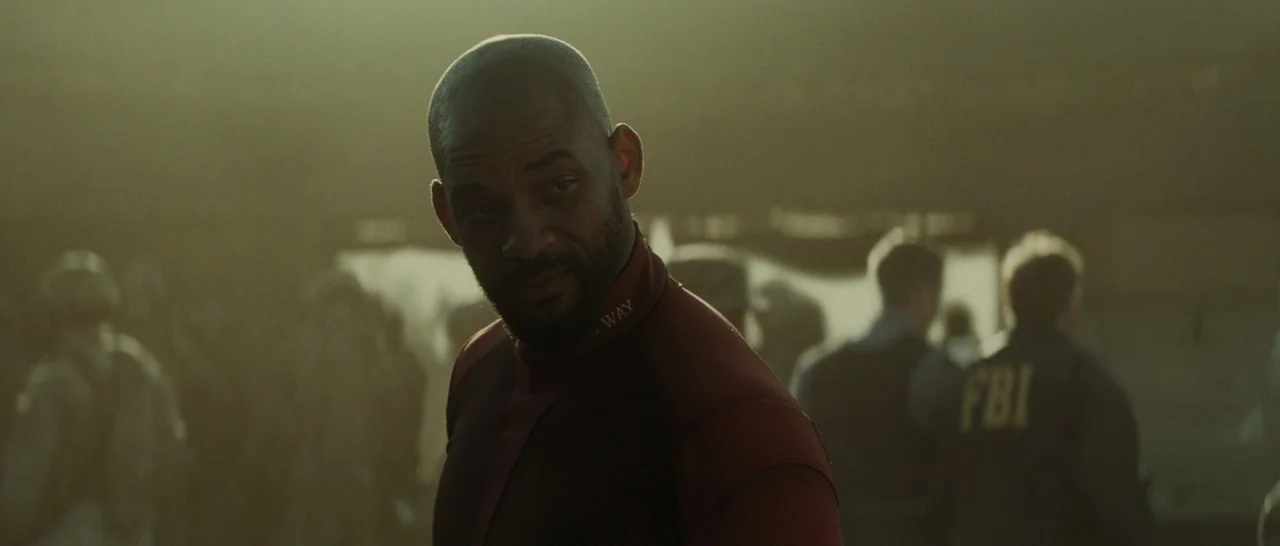 Suicide Squad (2016) movie still: medium — A hazy outdoor location filled with figures in uniform. A man in a maroon shirt stands in the foreground…