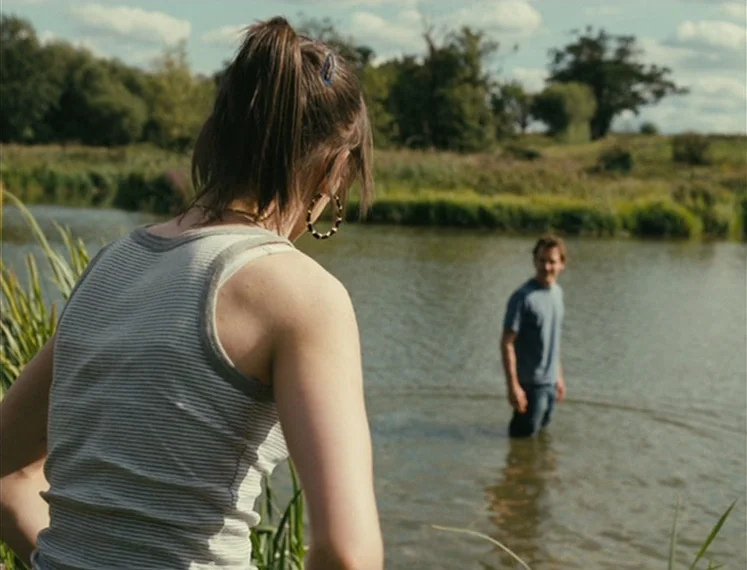 Fish Tank (2009) movie still: medium — A rural riverbank features tall grasses in the foreground and a line of trees. A man stands knee-deep in the…