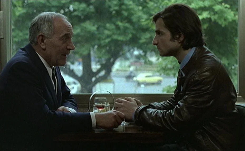 Love on the Run (1979) movie still: medium — Two men sit across from each other in a booth inside a restaurant. A window reveals a street with trees…