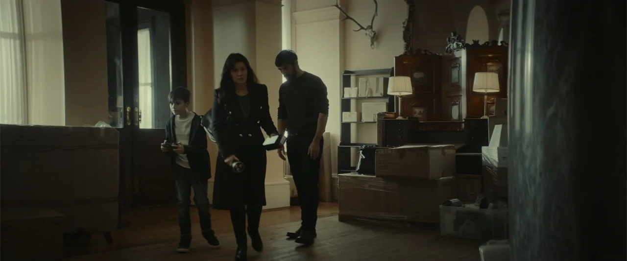 The Cellar (2022) movie still: fullbody — A living room filled with cardboard boxes indicates a recent move or transition. A woman, a man, and a boy…